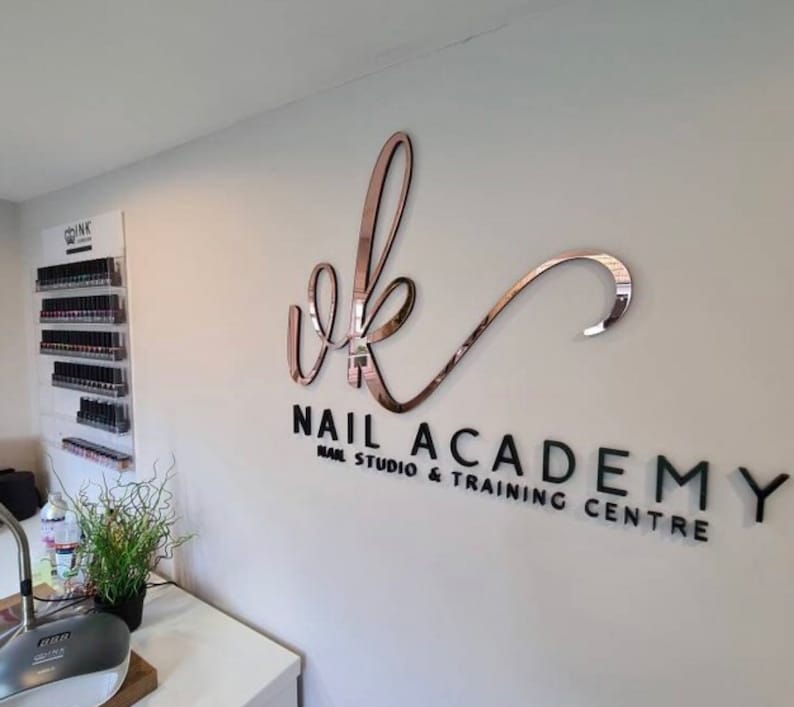 May include: A white wall with a black and rose gold sign that reads "vk Nail Academy Nail Studio & Training Centre".