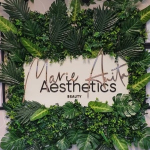 May include: A white sign with the text "Marie Aith Aesthetics Beauty" in a gold script font. The sign is surrounded by a green wall of artificial plants.