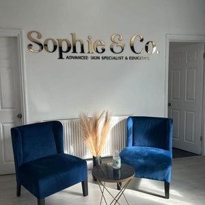 May include: A waiting area with two blue velvet chairs and a gold metal table with a black marble top. The chairs are facing each other and there is a vase with dried pampas grass on the table. The sign on the wall reads "Sophie & Co. Advanced Skin Specialist & Educator."