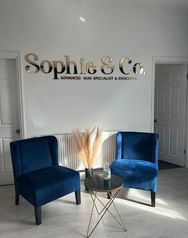 May include: A waiting area with two blue velvet chairs and a gold metal table with a black marble top. The chairs are facing each other and there is a vase with dried pampas grass on the table. The sign on the wall reads "Sophie & Co. Advanced Skin Specialist & Educator."