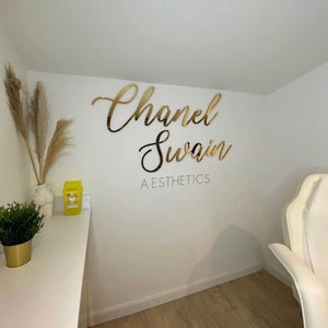 May include: Gold script lettering on a white wall reads "Chanel Swain AESTHETICS".