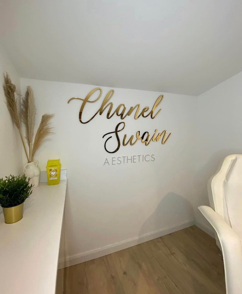 May include: Gold script lettering on a white wall reads "Chanel Swain AESTHETICS".