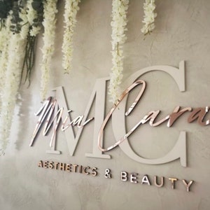 May include: A white wall with a rose gold sign that says "Mia Cara Aesthetics & Beauty". The sign is made of large, white letters with rose gold accents. The letters are in a cursive font. White flowers hang from the top of the image.