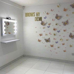 May include: A white vanity mirror with a row of light bulbs around the frame, a white shelf below the mirror, and a white wall with the text "Brows by Demi" and a cluster of pink, purple, and gold butterfly decorations.