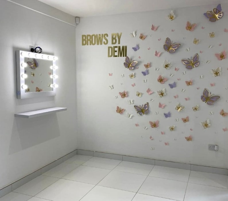 May include: A white vanity mirror with a row of light bulbs around the frame, a white shelf below the mirror, and a white wall with the text "Brows by Demi" and a cluster of pink, purple, and gold butterfly decorations.