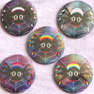 Pode incluir: Cinco botões redondos com pino com um fundo de galáxia e uma aranha preta com listras de diferentes cores nas suas pernas. As aranhas têm um corpo preto com olhos brancos e uma gravata borboleta preta. As listras nas pernas são nas cores das bandeiras do orgulho transgênero, bissexual, lésbica, gay e pansexual.