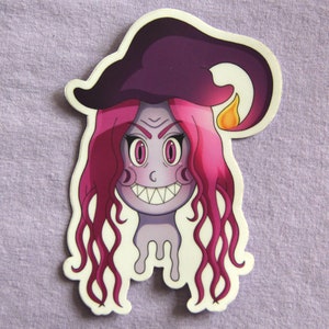 May include: A die-cut sticker featuring a cartoon witch with a purple face, pink hair, and a dark purple hat. The witch has a wide, toothy grin and a flame detail on the hat. The sticker has a white border and is set against a light purple background.