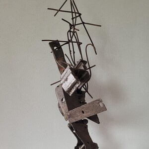 Pode incluir: Escultura abstrata em metal com uma mistura de formas geométricas e orgânicas. Apresenta uma base quadrada, peças de metal em camadas e finos elementos semelhantes a fios que se estendem para cima. A cor é marrom enferrujado.