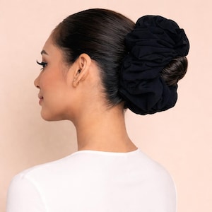 May include: A black scrunchie bun hairstyle. The scrunchie is large and made of a soft fabric. The hair is pulled back and styled in a bun. The person is wearing a white top.