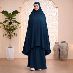 May include: A navy blue modest dress ensemble. The outfit includes a head covering, a long, flowing overdress, and a matching skirt. The ensemble is made of a soft, draping fabric. The background features a decorative window and neutral-toned vases.