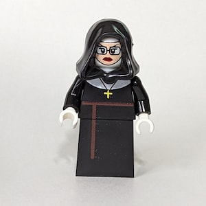 May include: A black Lego minifigure dressed as a nun. The figure has a black habit, a white wimple, and a gold cross necklace. The figure is wearing glasses and has a stern expression.