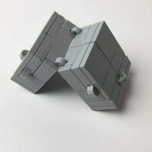 May include: A gray, articulated cube toy, composed of multiple rectangular blocks connected by small, circular hinges. The toy is positioned at an angle, showcasing its flexible design and geometric structure. The background is plain white.