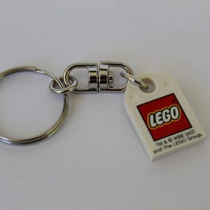 Może przedstawiać: Brelok do kluczy LEGO z białą zawieszką z logo LEGO w kolorze czerwonym i żółtym. Brelok posiada srebrny kółko do kluczy i obrotowy karabińczyk ułatwiający przypinanie do kluczy lub toreb.