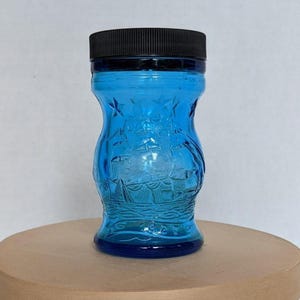 Puede incluir: Un tarro de vidrio azul con tapa negra. El tarro tiene un diseño en relieve de un velero y estrellas. El tarro es cilíndrico con una forma ligeramente de reloj de arena. El tarro está sobre una superficie marrón claro.