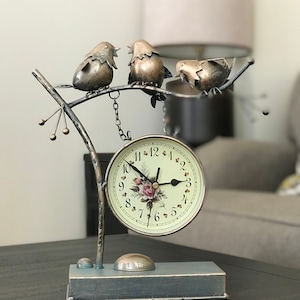 May include: A decorative clock with a metal bird design. The clock face has a floral pattern and the birds are perched on a branch. The clock is on a dark wood surface.