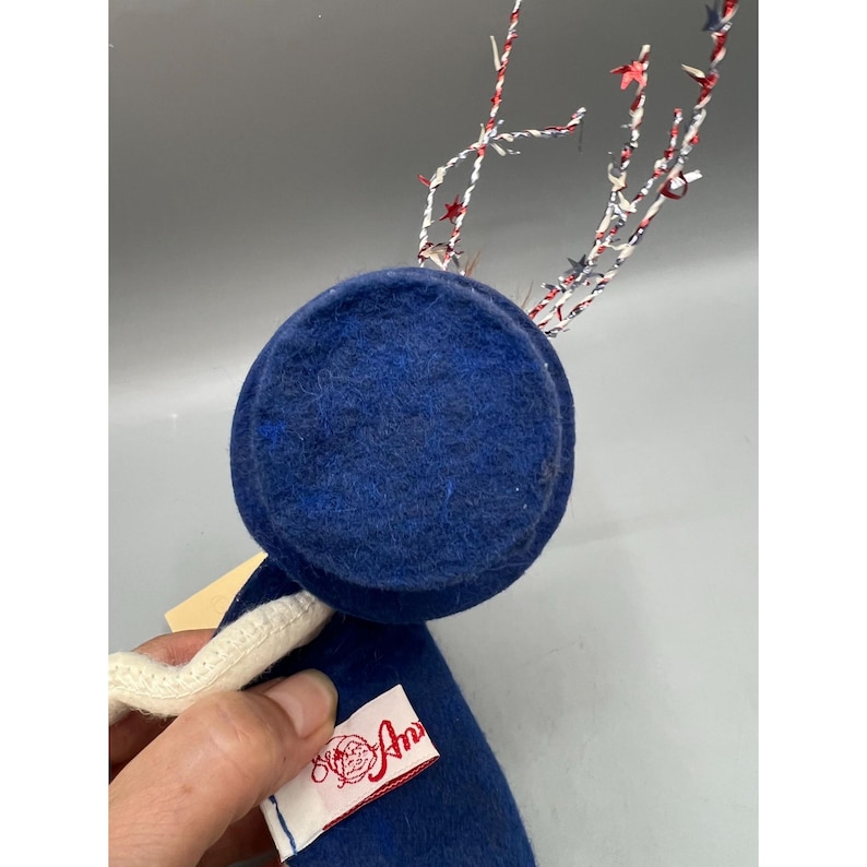 May include: A blue felt top hat ornament with a white trim and a tag reading Annalee. The hat features decorative wire antlers adorned with small red, white, and blue star-shaped accents, extending upward from the top of the blue felt surface.