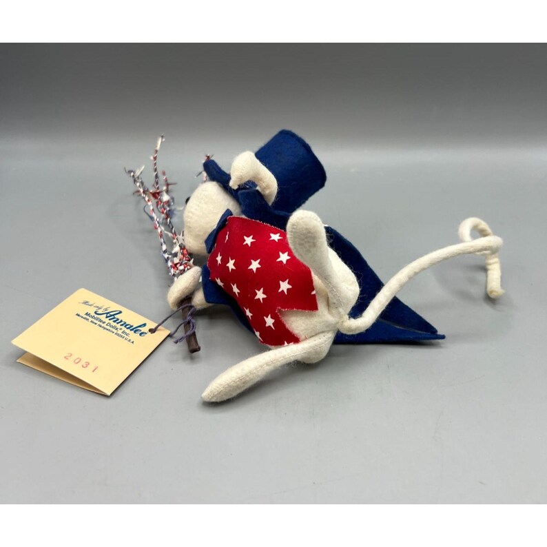 May include: A white felt mouse doll wearing a blue top hat and a red vest with white stars. The mouse holds a silver and red tinsel sprig. A cream-colored tag is attached, printed with "Annalee Mobilitee Dolls, Inc. Meredith, New Hampshire 03253 U.S.A." and "2031".