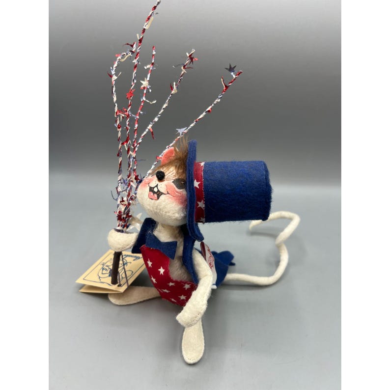 May include: A felt mouse figurine wearing a blue top hat with white stars, a blue bowtie, and a red apron with white stars. The mouse holds a bundle of red, white, and blue decorative twigs and a small rectangular paper tag with a line drawing.