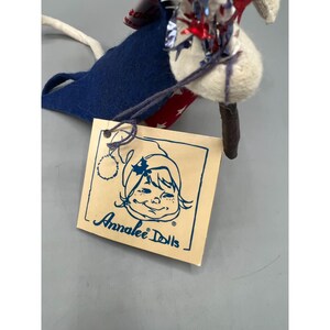 May include: A fabric doll featuring a blue felt body, a red and white star-patterned accent, and metallic red, white, and blue tinsel. A square beige tag is attached, displaying a line-art illustration of a face and the text Annalee Dolls.