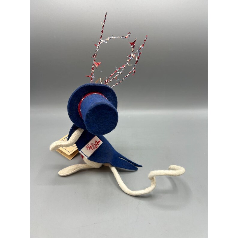 May include: A felt mouse figurine wearing a blue top hat and a matching blue tailcoat with a white lapel featuring the red text "Annalee". The mouse has a long white tail and is adorned with decorative red, white, and blue tinsel-like wire antlers.