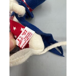 May include: A felt doll wearing a blue hat and a red cape with white stars. A white fabric tag is attached, featuring red text that reads: "alee '97 MADE ONLY IN MEREDITH NEW HAMPSHIRE, U.S.A." The doll has a white tail and blue felt accents.
