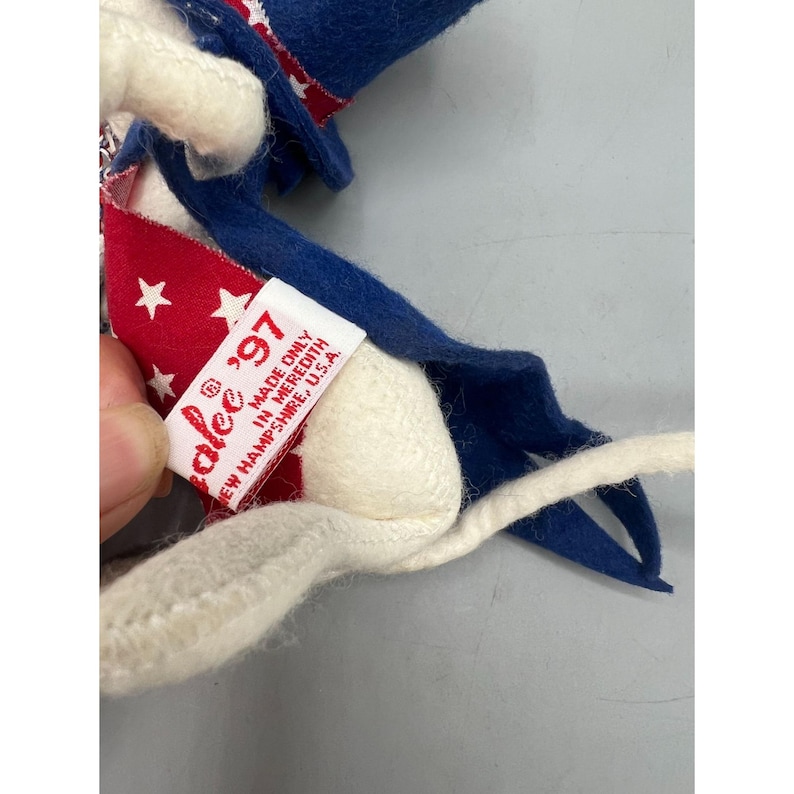 May include: A felt doll wearing a blue hat and a red cape with white stars. A white fabric tag is attached, featuring red text that reads: "alee '97 MADE ONLY IN MEREDITH NEW HAMPSHIRE, U.S.A." The doll has a white tail and blue felt accents.