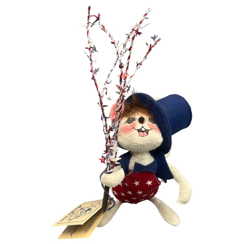May include: A plush mouse figurine wearing a dark blue top hat, a matching vest, and red shorts with white stars. The mouse holds a tall, thin branch decorated with red, white, and blue star-shaped tinsel. A paper tag is attached to the base.