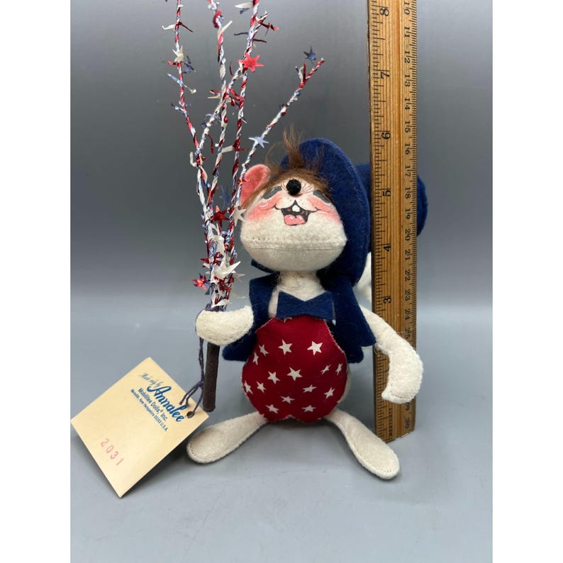 May include: A fabric mouse doll wearing a blue hat, blue vest, and red overalls with white stars. The mouse holds a decorative branch with red, white, and blue star accents. A wooden ruler shows the doll is approximately 9 inches tall.