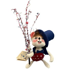 May include: A felt mouse figurine wearing a blue top hat, a blue bowtie, and red overalls with white stars. The mouse has brown fur on its head, pink cheeks, and holds a decorative branch adorned with red, white, and blue star-shaped accents.