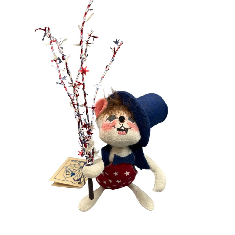 May include: A felt mouse figurine wearing a blue top hat, a blue bowtie, and red overalls with white stars. The mouse has brown fur on its head, pink cheeks, and holds a decorative branch adorned with red, white, and blue star-shaped accents.