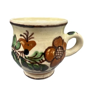 May include: A small, off-white ceramic mug with a handle, adorned with hand-painted floral and leaf designs in brown, green, and black. The mug features a rounded base and a slightly flared rim, showcasing a rustic, folk art aesthetic.