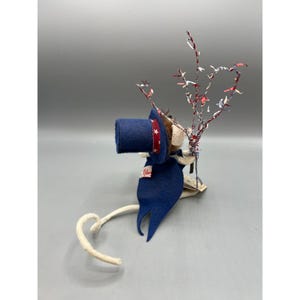 May include: A felt mouse figurine wearing a blue top hat with a red and white star-patterned band and a matching blue cape. The mouse holds a decorative branch adorned with small red, white, and blue star-shaped confetti pieces.