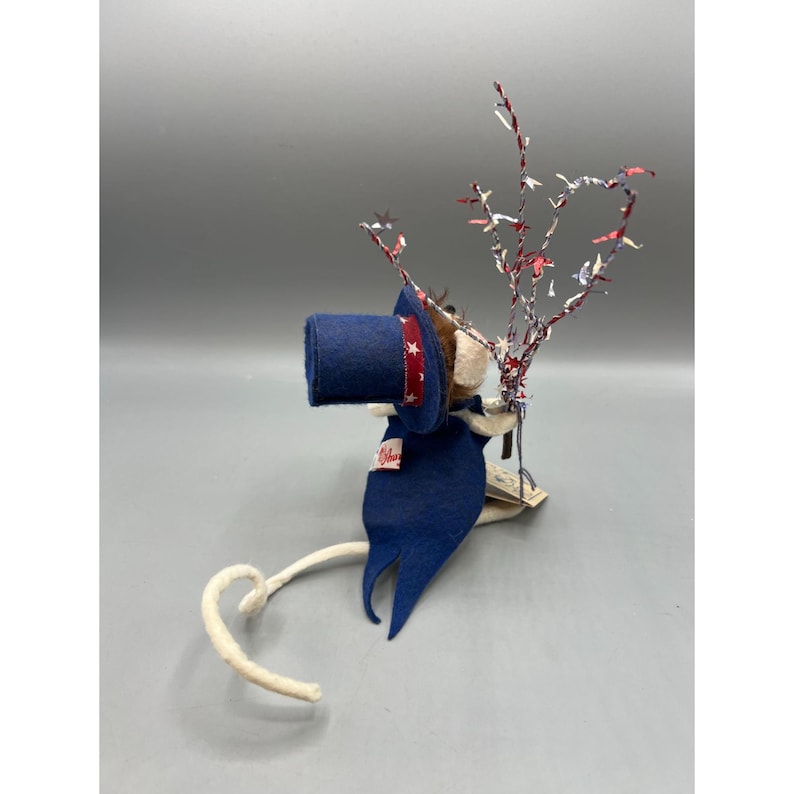 May include: A felt mouse figurine wearing a blue top hat with a red and white star-patterned band and a matching blue cape. The mouse holds a decorative branch adorned with small red, white, and blue star-shaped confetti pieces.