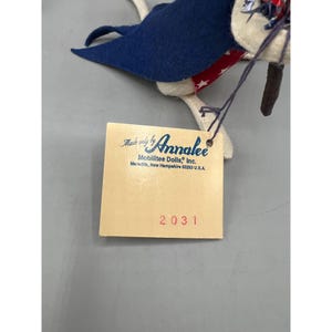 May include: A felt doll featuring a blue cape and a red fabric trim with white stars. A square cream-colored tag is attached, printed with Made only by Annalee Mobilitee Dolls, Inc. Meredith, New Hampshire 03253 U.S.A. and the red number 2031.