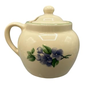 May include: Cream-colored ceramic sugar bowl with a lid and handle. The bowl is decorated with a hand-painted design of blue blueberries and green leaves. The lid has a small knob on top.