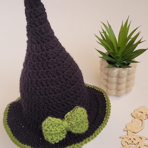 May include: A black crochet witch hat with a green trim and bow. This handmade hat is a Halloween decoration. The hat is next to a small potted plant. The hat is a fun accessory for costumes.