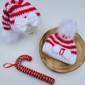 May include: Two crochet Santa hats with white bows and faux fur pom-poms. One is tall, the other a beanie. A red and white glitter candy cane ornament is also in the image. The hats have red and white stripes.