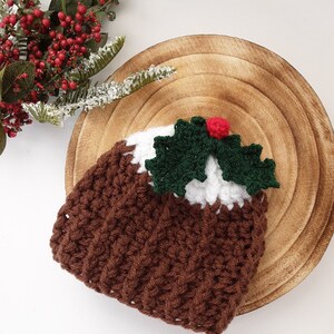 May include: A crocheted Christmas pudding, featuring brown, white, and green elements, and a red berry on top. It rests on a wooden plate, with a sprig of red berries and greenery in the background. This handmade item is a festive decoration for the holiday season.