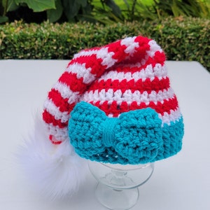 May include: A handmade crochet Santa hat with red and white stripes, a turquoise bow, and a white fluffy pom-pom. The hat is displayed on a clear glass pedestal, perfect for holiday celebrations.