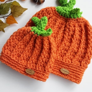 May include: Two crocheted pumpkin hats, one larger than the other, with green crocheted stems. The smaller hat has a wooden tag that says "Little Pumpkin" and the larger hat has a wooden tag that says "Pumpkin".