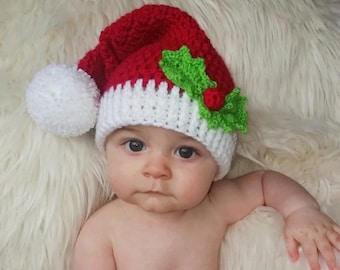 May include: A red and white crocheted Santa hat with a white pom-pom and green holly leaves with a red berry. The hat is worn by a baby. The hat is a festive accessory for the holiday season.