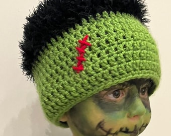 May include: Green and black crocheted hat with a black fuzzy top and red stitching. The hat is worn by a child with green face paint and stitches.