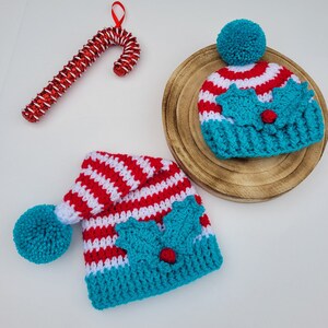 May include: Two crocheted Christmas hats with red and white stripes, teal accents, and holly leaf details. One hat has a teal pom-pom. A red and white glitter candy cane ornament is also present.