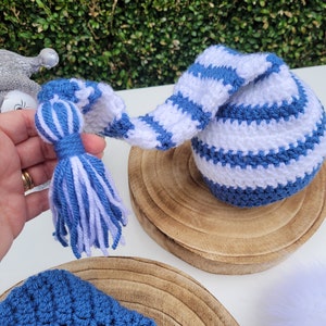 May include: A handmade crochet gnome hat in blue and white stripes. The hat has a long, curved tail with a tassel at the end. The hat is sitting on a wooden surface. The hat is a festive item.