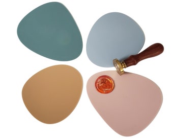May include: Four colorful, irregularly shaped silicone coasters in blue, light blue, beige, and pink. A wooden handle with a gold wax seal is on top of the pink coaster.