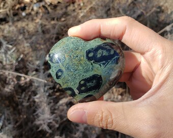 May include: A green and black heart-shaped stone with a swirling pattern. The stone is being held in a hand.