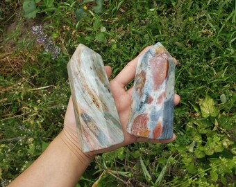 May include: Two colorful, pointed, natural stone crystals. The crystals are a mix of white, gray, brown, red, and blue. The crystals are held in a hand.