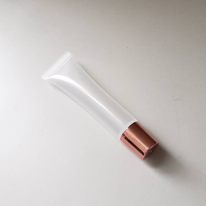 May include: A white, empty squeeze tube with a metallic rose gold cap. The tube is made of a translucent material, allowing the contents to be visible. The cap is a solid, shiny color. The tube is angled on a white surface.