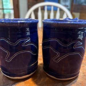May include: Two handmade ceramic mugs. The mugs are a deep blue with a lighter blue rim and a white, wave-like design. The mugs are sitting on a wooden surface.