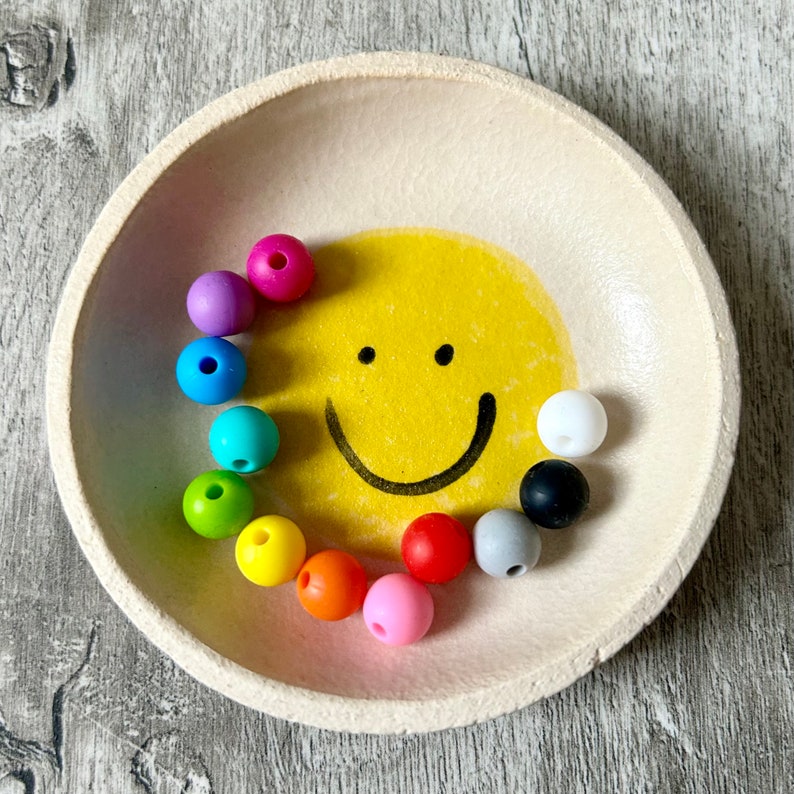 Op de afbeelding: Een witte keramische kom met een geel smiley-gezicht geverfd op de bodem. De kom is gevuld met kleurrijke siliconen kralen in verschillende tinten rood, oranje, geel, groen, blauw en paars.